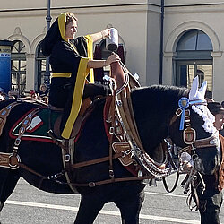 „Münchner Kindl“ mit schwarzem und gelbem Gewand reitet auf geschmücktem Pferd bei Trachten- und Schützenzug vor Zuschauergruppe