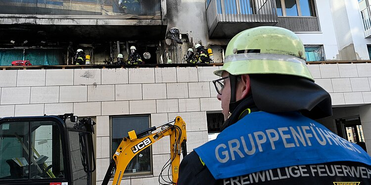 Feuerwehrmann mit Helm und 'Gruppenführer Freiwillige Feuerwehr München' auf der Jacke vor einem ausgebrannten Wohngebäude mit Feuerwehrleuten auf dem Balkon