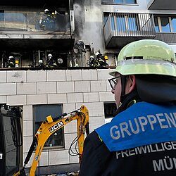 Feuerwehrmann mit Helm und 'Gruppenführer Freiwillige Feuerwehr München' auf der Jacke vor einem ausgebrannten Wohngebäude mit Feuerwehrleuten auf dem Balkon