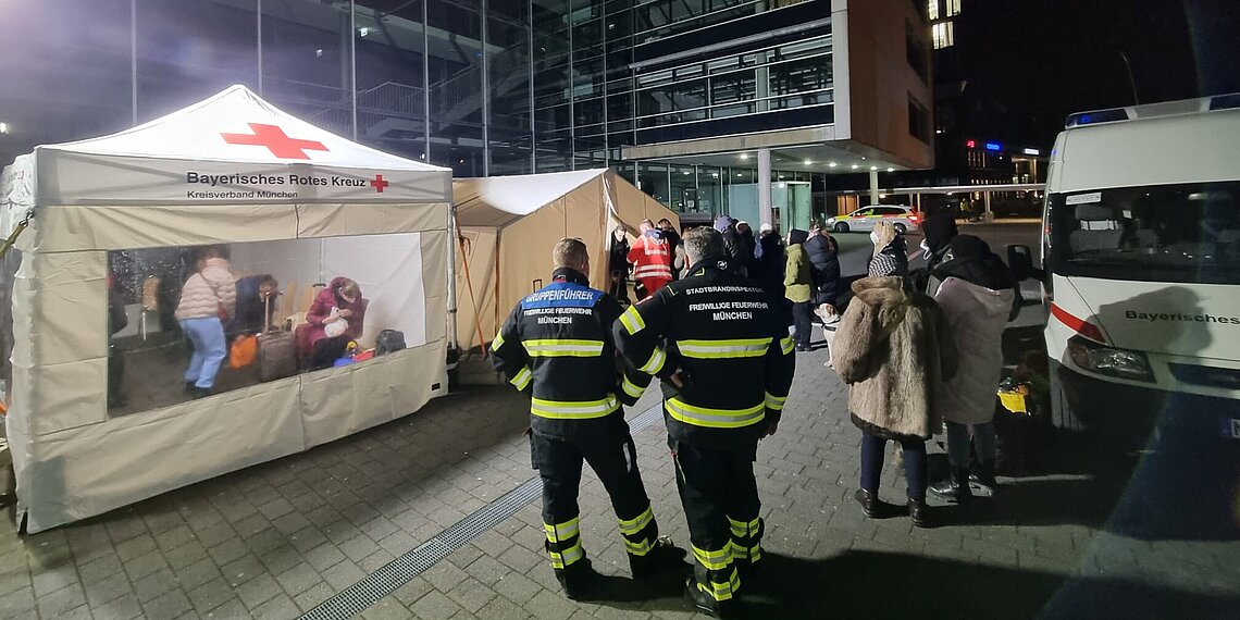 Zelt des BRK bei Ukraine-Hilfe-Einsatz mit Einsatzkräften und Menschen in der Nacht vor einem Gebäude
