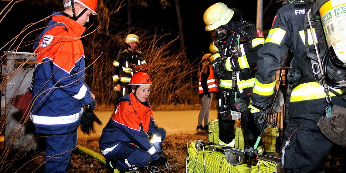 Feuerwehrleute der Jugendfeuerwehr und erwachsene Einsatzkräfte in Schutzkleidung und Helmen bei nächtlichem Übungseinsatz mit Schlauchtragekorb und Atemschutzgeräten