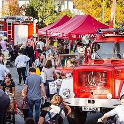 Menschenmenge beim Tag der offenen Tür der Abteilung Harthof mit roten Feuerwehrfahrzeugen und Pavillons in einer Straße mit herbstlichen Bäumen