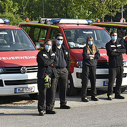 Feuerwehrleute in schwarzer Uniform und Mundschutz stehen vor Feuerwehrfahrzeugen bei einer Masken-Verteilaktion im Freien
