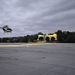 Polizeihubschrauber im Schwebeflug über der Theresienwiese bei Dämmerung, Flughelfer und Einsatzfahrzeuge sind vor Ort, im Hintergrund die Bavaria