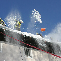 Mehrere Feuerwehrleute räumen Schnee mit Schaufeln von einem Dach, Schnee fliegt in die Luft, Szene bei klarem Himmel