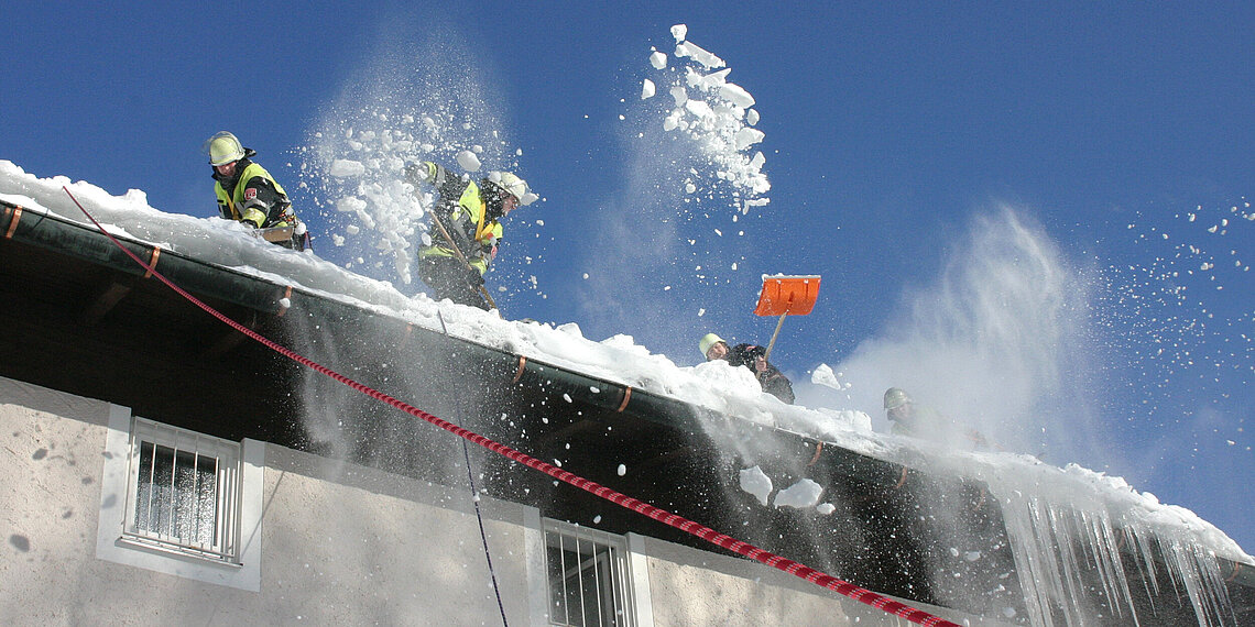 Mehrere Feuerwehrleute räumen Schnee mit Schaufeln von einem Dach, Schnee fliegt in die Luft, Szene bei klarem Himmel