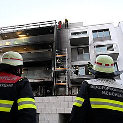 Feuerwehrleute der Feuerwehr München beobachten einen Einsatz an einem mehrstöckigen Wohngebäude mit beschädigter Fassade und einer Leiter an der Außenwand