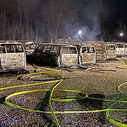 Ausgebrannte Polizeifahrzeuge stehen nachts auf einem Schotterplatz, umgeben von Löschschläuchen der Feuerwehr