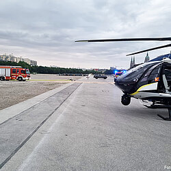 Polizeihubschrauber auf der Theresienwiese mit einem Feuerwehrfahrzeug und mehreren Einsatzfahrzeugen im Hintergrund unter bewölktem Himmel