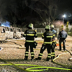 Ausgebrannte Polizeifahrzeuge nachts mit Einsatzkräften der Feuerwehr mit Löschschläuchen