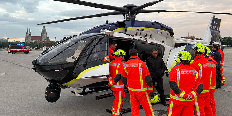 Polizeihubschrauber Airbus H145 auf der Theresienwiese, umgeben von Flughelfern in leuchtend orangen Schutzanzügen und Helmen, Einsatzfahrzeug im Hintergrund