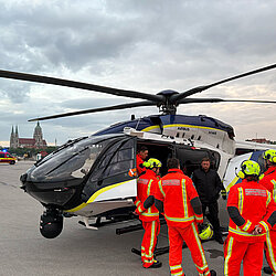 Polizeihubschrauber Airbus H145 auf der Theresienwiese, umgeben von Flughelfern in leuchtend orangen Schutzanzügen und Helmen, Einsatzfahrzeug im Hintergrund