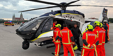 Polizeihubschrauber Airbus H145 auf der Theresienwiese, umgeben von Flughelfern in leuchtend orangen Schutzanzügen und Helmen, Einsatzfahrzeug im Hintergrund