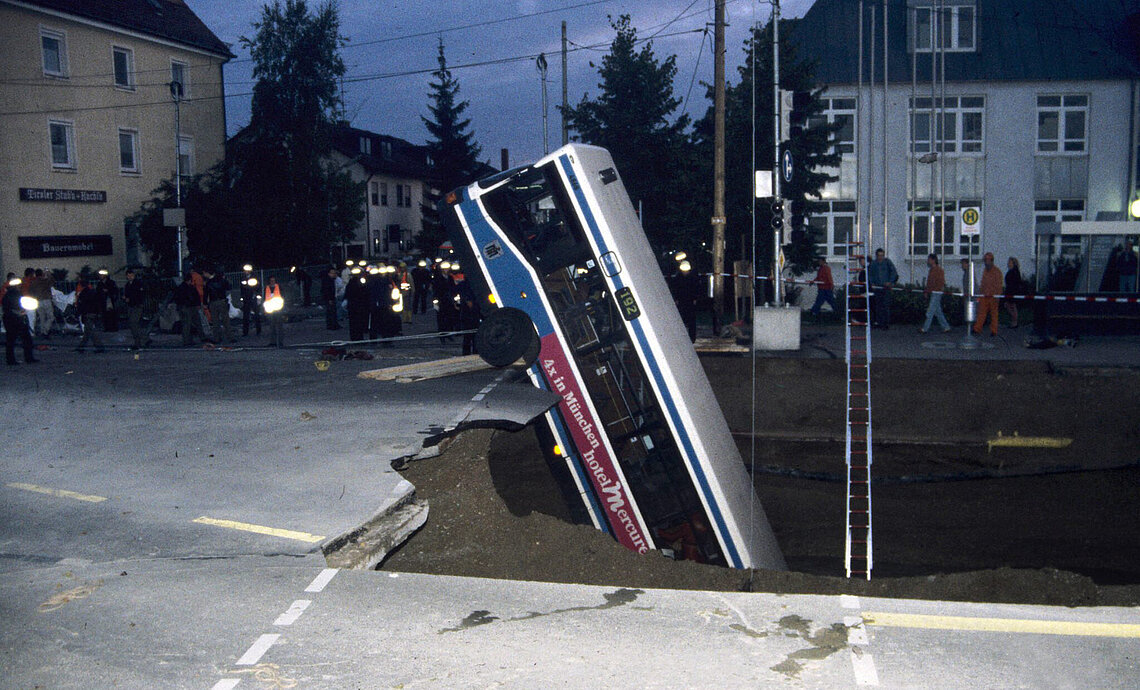 Omnibus in Trudering nach Busunglück steckt bei Dämmerung schräg in großem Straßeneinbruch, Einsatzkräfte und Zuschauer im Hintergrund