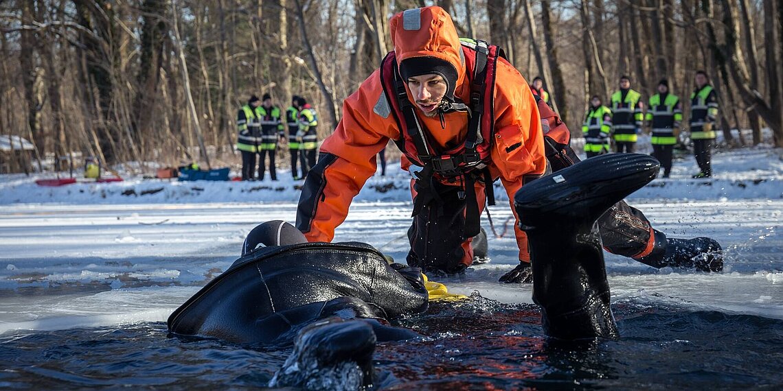 Rettungskraft in orangenem Schutzanzug zieht eine ins Eis eingebrochene Person aus einem zugefrorenen Gewässer, im Hintergrund weitere Einsatzkräfte an der Eisfläche