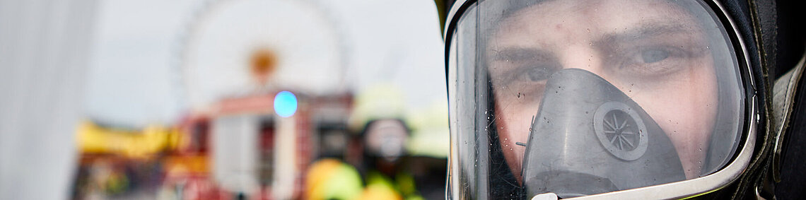 Nahaufnahme eines Feuerwehrmanns mit Atemschutzmaske und Helm, unscharfer Einsatzhintergrund mit Feuerwehrfahrzeug Nahaufnahme eines Feuerwehrmanns mit Atemschutzmaske und Helm, unscharfer Einsatzhintergrund mit Feuerwehrfahrzeug