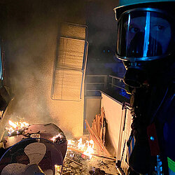 Feuerwehrmann mit Helm und Atemschutzmaske steht bei Dunkelheit neben Unrat auf einem Balkon, der dort brennt