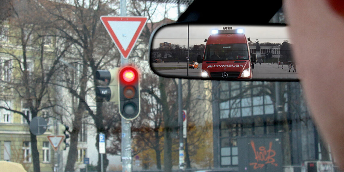 Im Rückspiegel zu sehen ist ein Einsatzfahrzeug der Feuerwehr mit Blaulicht, während die Ampel vor dem Fahrer rot zeigt