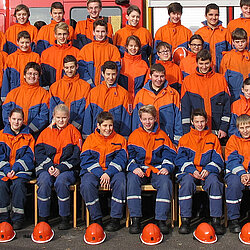 Gruppenfoto der Jugendfeuerwehr München mit Jugendlichen in orange-blauen Uniformen und Ausbildern in schwarzer Schutzkleidung vor und auf einem Feuerwehrfahrzeug