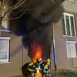 Zwei Feuerwehrleute in Schutzkleidung löschen ein Feuer in einem Hauseingang eines Wohngebäudes, aus dem dichter schwarzer Rauch und Flammen aufsteigen