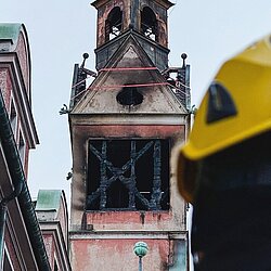 Verbrannter Kirchturm mit beschädigten Fenstern, Einsatzkraft mit gelbem Helm im Vordergrund