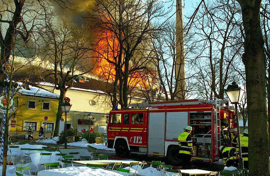 Feuerwehrfahrzeug und Einsatzkräfte löschen Brand im Salvatorkeller-Gebäude, Flammen und Rauch steigen aus dem Dach auf, Tische und Stühle im Vordergrund