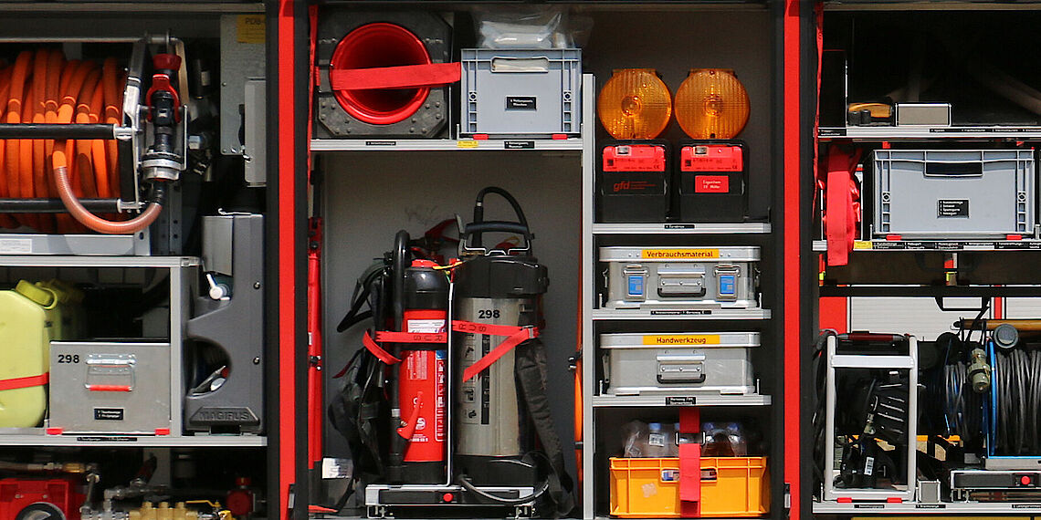 Feuerwehrfahrzeug mit geöffnetem Geräteraum, zeigt verschiedene Geräte wie Feuerlöscher, Signalleuchten, Werkzeugkisten und Kabel
